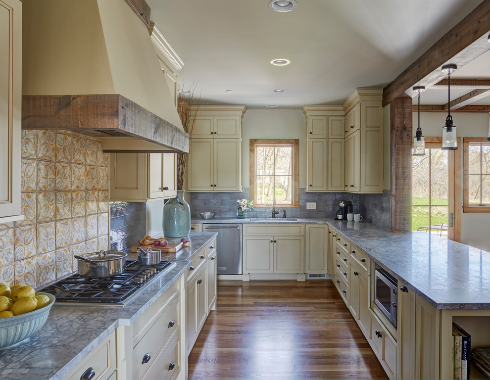 Mid-sized country u-shaped eat-in kitchen in Chicago with a single-bowl sink, recessed-panel cabinets, beige cabinets, quartzite benchtops, grey splashback, cement tile splashback, stainless steel appliances, light hardwood floors, a peninsula, brown floor, grey benchtop and exposed beam.