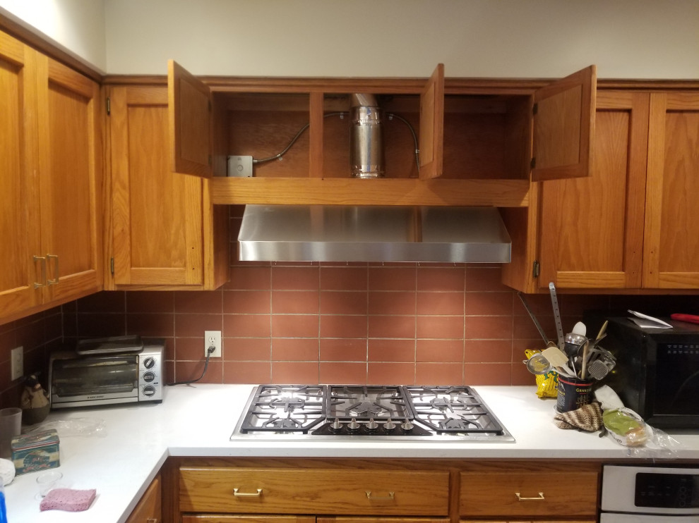This is an example of a large country l-shaped eat-in kitchen in Kansas City with an undermount sink, shaker cabinets, light wood cabinets, granite benchtops, red splashback, terra-cotta splashback, stainless steel appliances, light hardwood floors, with island, brown floor and white benchtop.