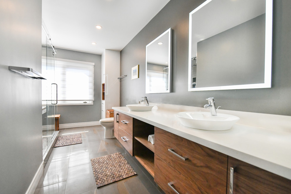 This is an example of a large modern 3/4 bathroom in Toronto with flat-panel cabinets, medium wood cabinets, a corner shower, gray tile, porcelain tile, grey walls, concrete floors, a vessel sink, solid surface benchtops, grey floor, a sliding shower screen and white benchtops.