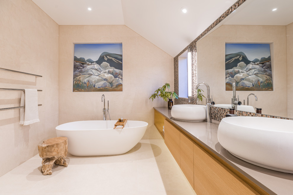 This is an example of a large contemporary master bathroom in Auckland with flat-panel cabinets, a freestanding tub, beige tile, limestone, beige walls, limestone floors, a vessel sink, engineered quartz benchtops, beige floor, medium wood cabinets and grey benchtops.
