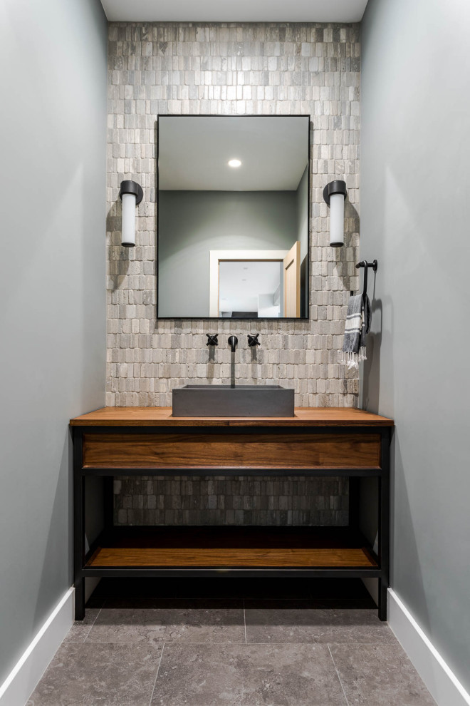 Inspiration for a mid-sized transitional powder room in Denver with medium wood cabinets, a two-piece toilet, multi-coloured tile, limestone, grey walls, porcelain floors, a vessel sink, wood benchtops, grey floor, brown benchtops and a freestanding vanity.