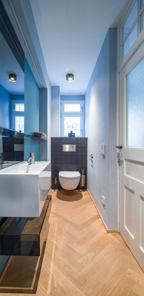 Mid-sized contemporary powder room in Stuttgart with a wall-mount toilet, gray tile, ceramic tile, blue walls, an integrated sink, light hardwood floors and brown floor.
