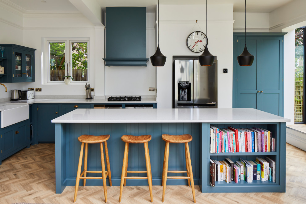 Design ideas for a mid-sized transitional l-shaped kitchen in London with a farmhouse sink, shaker cabinets, blue cabinets, stainless steel appliances, light hardwood floors, with island, beige floor and white benchtop.