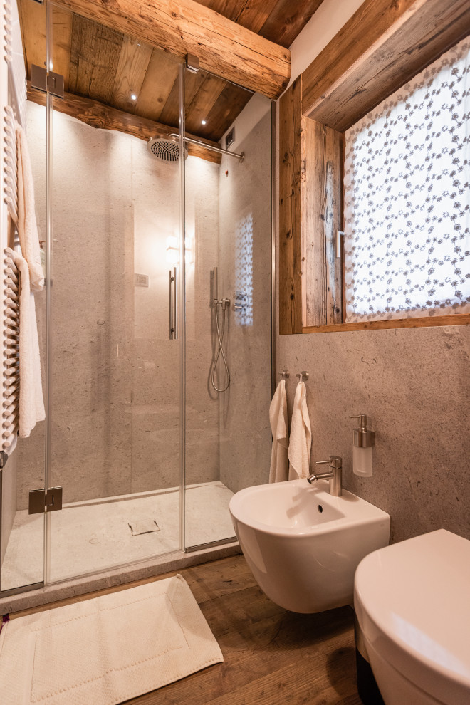 Small country 3/4 bathroom with recessed-panel cabinets, medium wood cabinets, a corner shower, a wall-mount toilet, gray tile, limestone, white walls, medium hardwood floors, a vessel sink, wood benchtops and a hinged shower door.