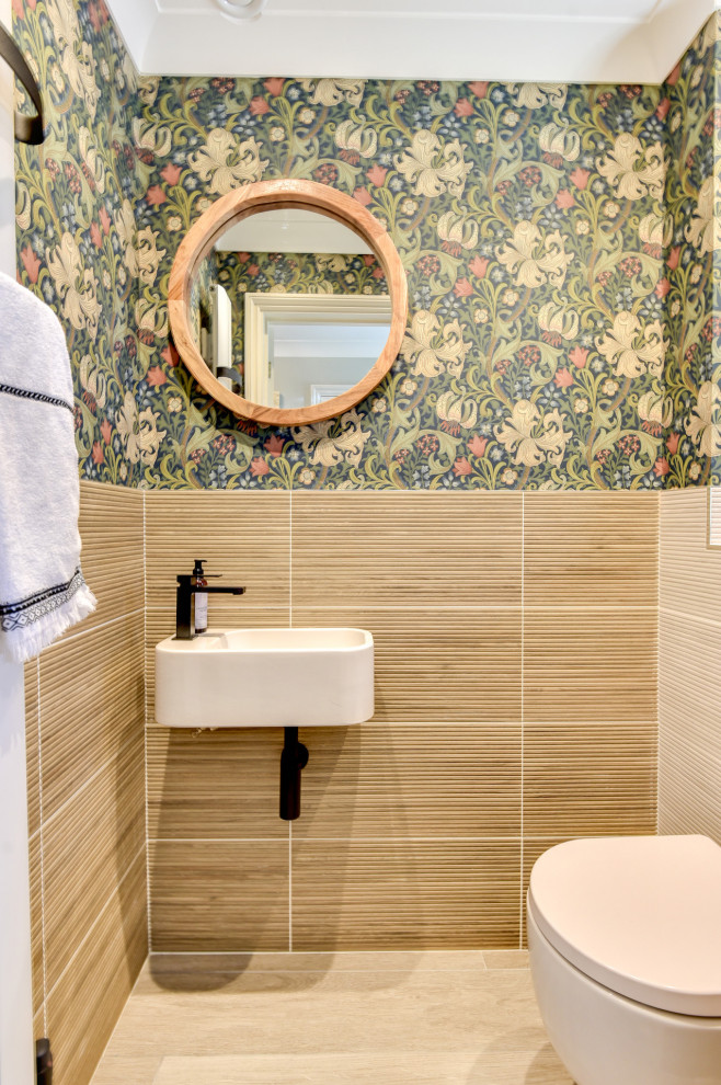 This is an example of a small modern powder room in Sussex with a one-piece toilet, beige tile, ceramic tile, multi-coloured walls, ceramic floors, a wall-mount sink, tile benchtops, beige floor, beige benchtops and wallpaper.