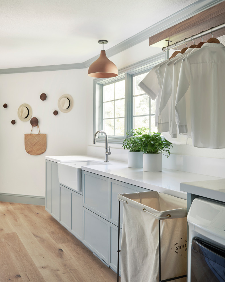 Photo of a large modern galley utility room in San Francisco with shaker cabinets, a farmhouse sink, grey cabinets, quartz benchtops, white splashback, engineered quartz splashback, white walls, light hardwood floors, a side-by-side washer and dryer and white benchtop.