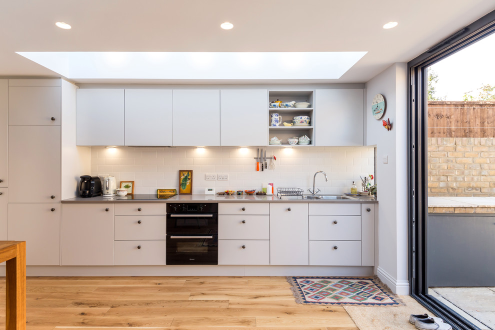 This is an example of a mid-sized contemporary single-wall eat-in kitchen in London with a single-bowl sink, flat-panel cabinets, white cabinets, white splashback, subway tile splashback, light hardwood floors, beige floor, grey benchtop, quartzite benchtops, black appliances and no island.