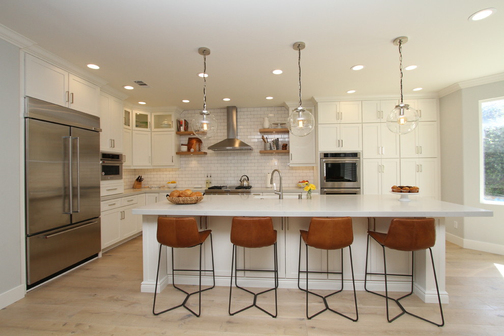Photo of a large transitional l-shaped open plan kitchen in Orange County with a farmhouse sink, shaker cabinets, white cabinets, quartz benchtops, white splashback, subway tile splashback, stainless steel appliances, light hardwood floors, with island and beige floor.
