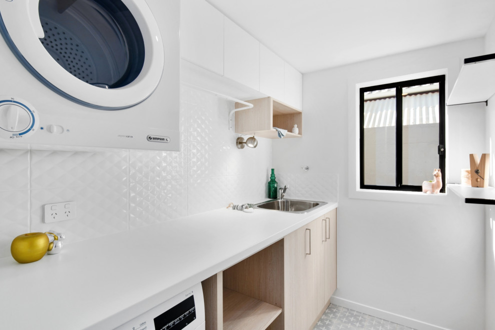 Photo of a small modern single-wall dedicated laundry room in Brisbane with a single-bowl sink, light wood cabinets, laminate benchtops, white splashback, ceramic splashback, white walls, ceramic floors, a stacked washer and dryer, multi-coloured floor and white benchtop.