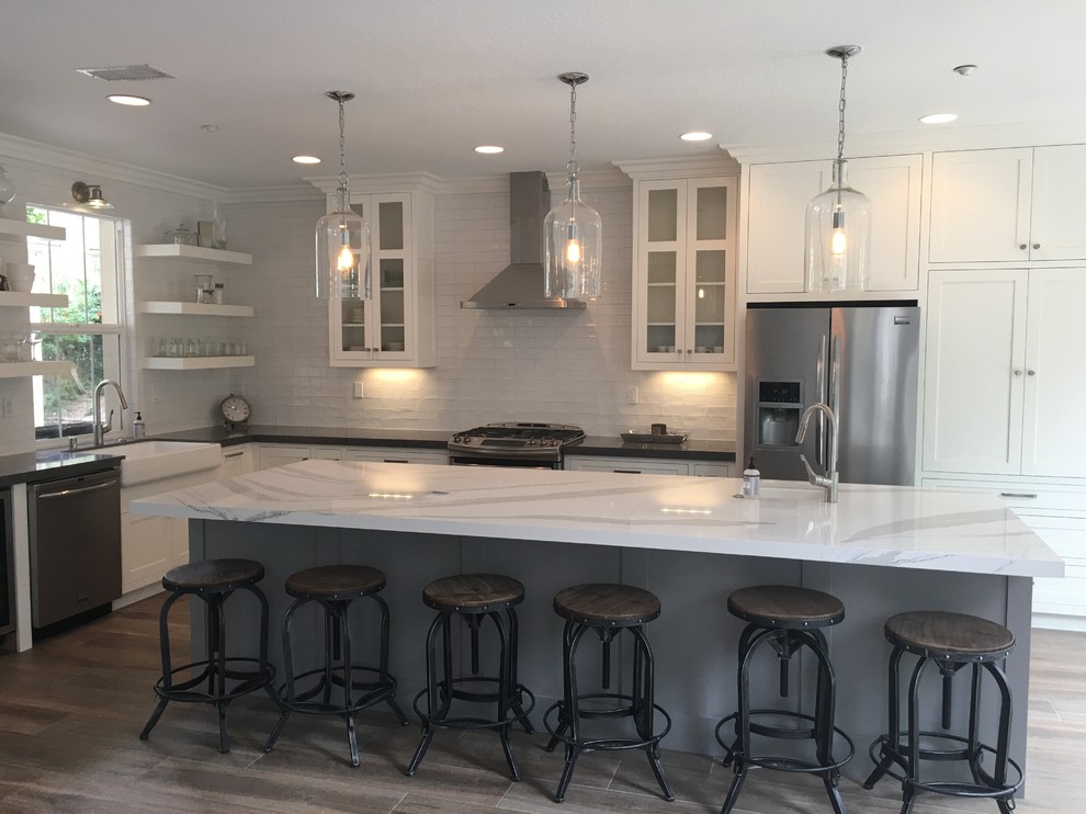 Inspiration for a mid-sized transitional l-shaped medium tone wood floor eat-in kitchen remodel in Orange County with a farmhouse sink, shaker cabinets, white cabinets, marble countertops, white backsplash, brick backsplash, stainless steel appliances and an island