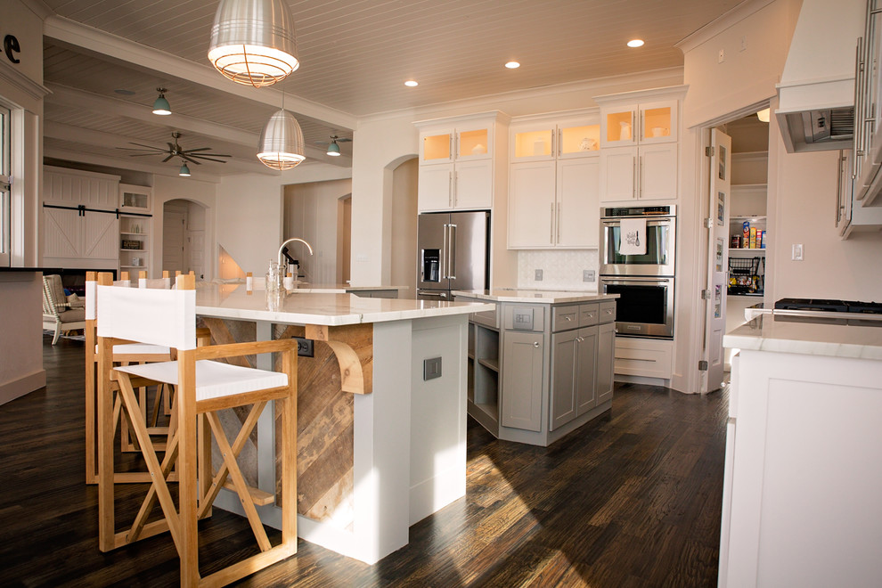 Inspiration for a large beach style l-shaped open plan kitchen in Dallas with a farmhouse sink, shaker cabinets, white cabinets, marble benchtops, white splashback, ceramic splashback, stainless steel appliances, dark hardwood floors, multiple islands and brown floor.