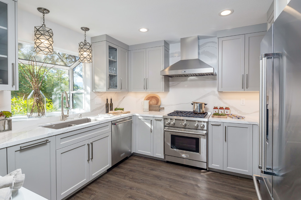 This is an example of a mid-sized transitional u-shaped kitchen in San Francisco with quartz benchtops, white splashback, stainless steel appliances, medium hardwood floors, no island, brown floor, white benchtop, a double-bowl sink, shaker cabinets and grey cabinets.