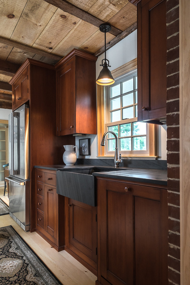 Photo of a small country l-shaped separate kitchen in Portland Maine with a farmhouse sink, stainless steel appliances, light hardwood floors, a peninsula, recessed-panel cabinets, dark wood cabinets, soapstone benchtops and red splashback.