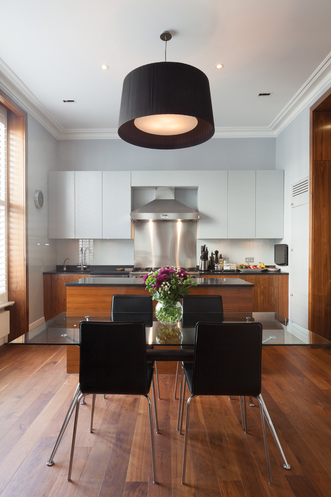 Photo of a large eclectic galley eat-in kitchen in London with a drop-in sink, flat-panel cabinets, light wood cabinets, granite benchtops, blue splashback, glass sheet splashback, panelled appliances, medium hardwood floors, with island, brown floor and black benchtop.