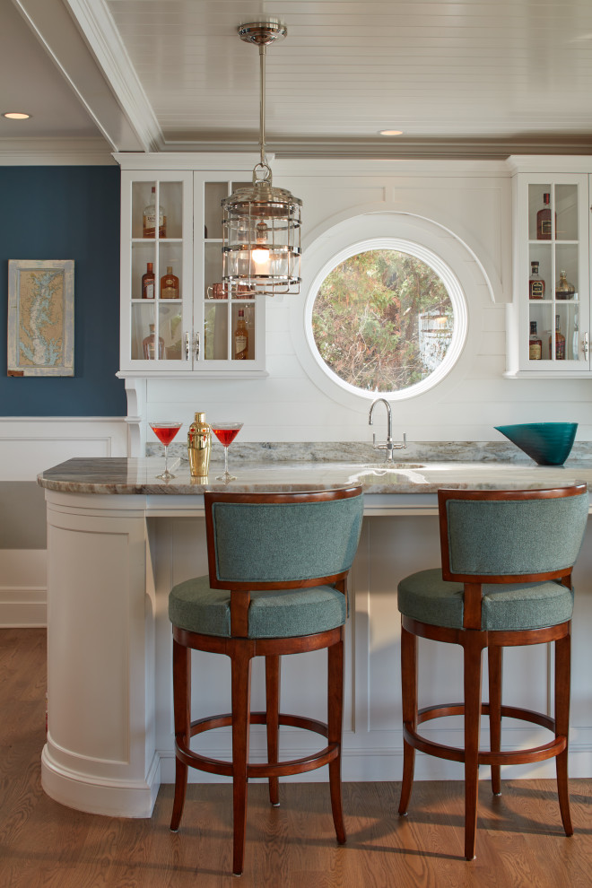 Photo of a beach style single-wall seated home bar in Baltimore with an integrated sink, glass-front cabinets, white cabinets, white splashback, shiplap splashback, medium hardwood floors, brown floor and grey benchtop.