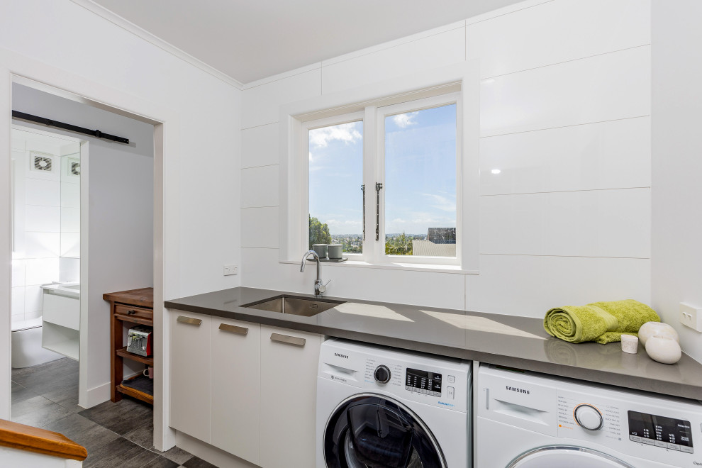 Mid-sized modern single-wall dedicated laundry room in Auckland with an undermount sink, flat-panel cabinets, white cabinets, solid surface benchtops, white splashback, ceramic splashback, white walls, porcelain floors, a side-by-side washer and dryer, grey floor and grey benchtop.