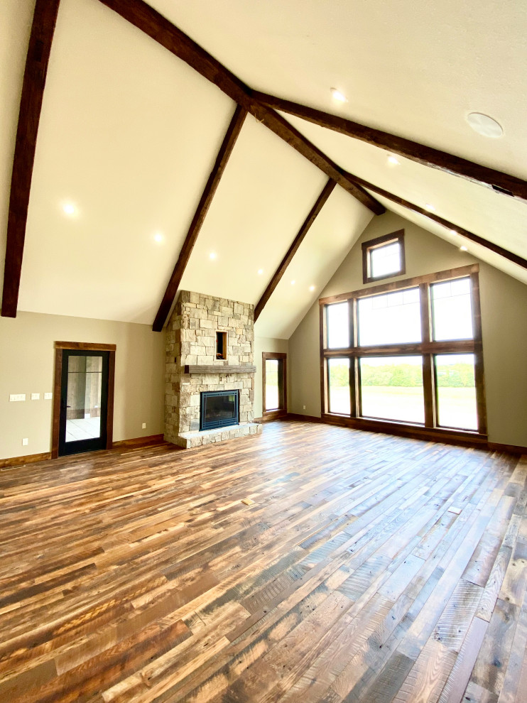 This is an example of a large rustic open plan games room in Kansas City with brown walls, medium hardwood flooring, a stone fireplace surround, a wall mounted tv, brown floors and exposed beams.
