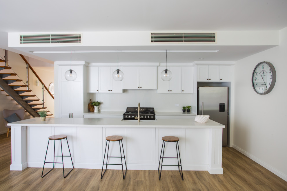 Design ideas for a large contemporary galley eat-in kitchen in Brisbane with a farmhouse sink, shaker cabinets, white cabinets, quartz benchtops, white splashback, engineered quartz splashback, black appliances, medium hardwood floors, with island, brown floor and white benchtop.