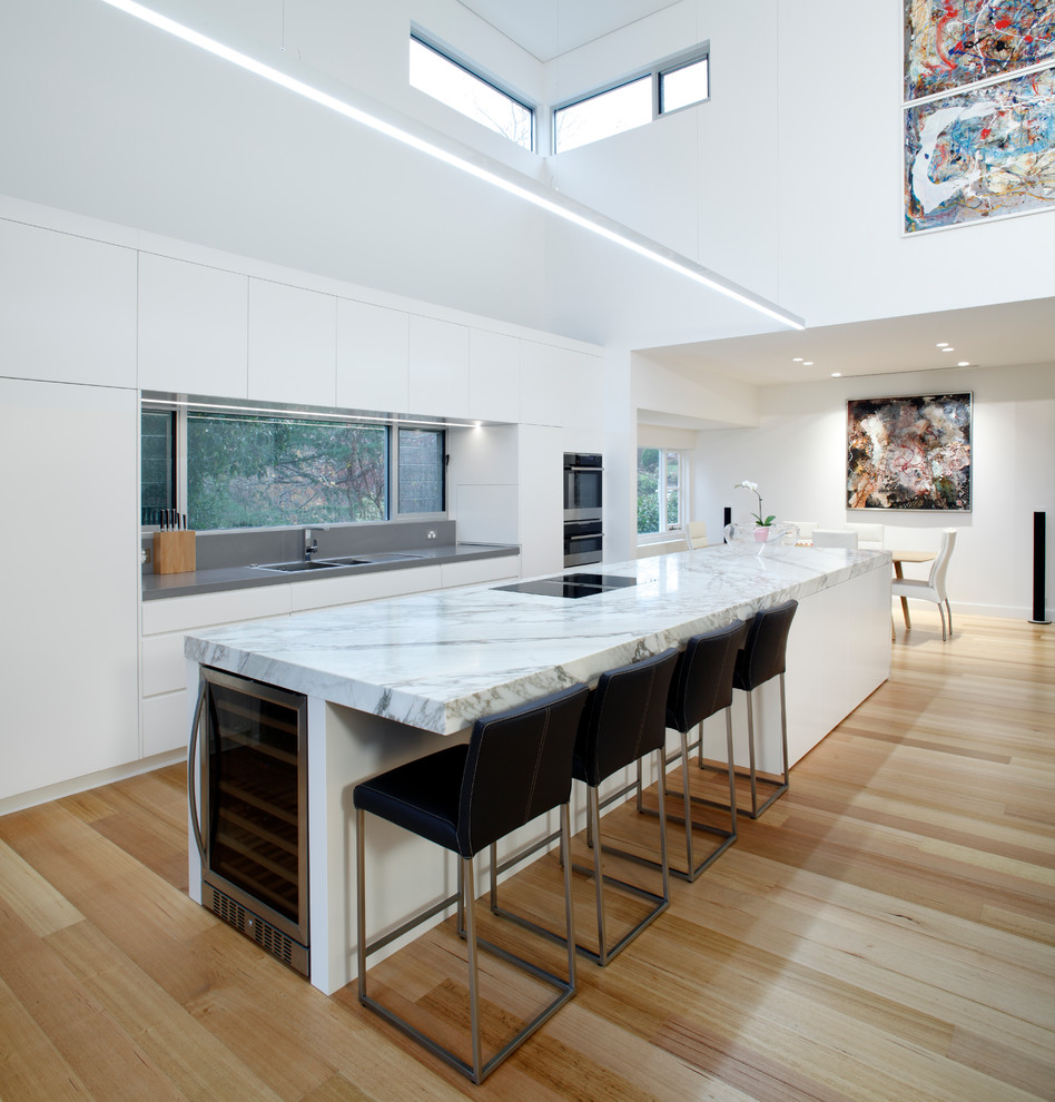 Design ideas for a mid-sized contemporary galley open plan kitchen in Wollongong with a double-bowl sink, flat-panel cabinets, white cabinets, marble benchtops, stainless steel appliances, light hardwood floors and with island.
