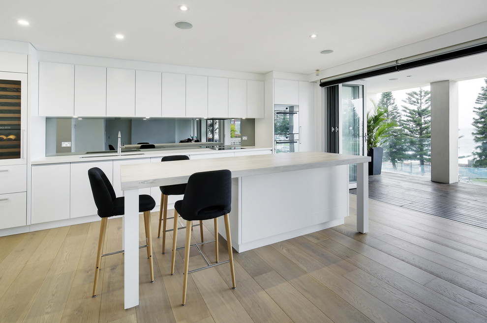 This is an example of a large scandinavian l-shaped open plan kitchen in Sydney with an undermount sink, flat-panel cabinets, white cabinets, quartz benchtops, metallic splashback, mirror splashback, black appliances, light hardwood floors, with island and yellow floor.