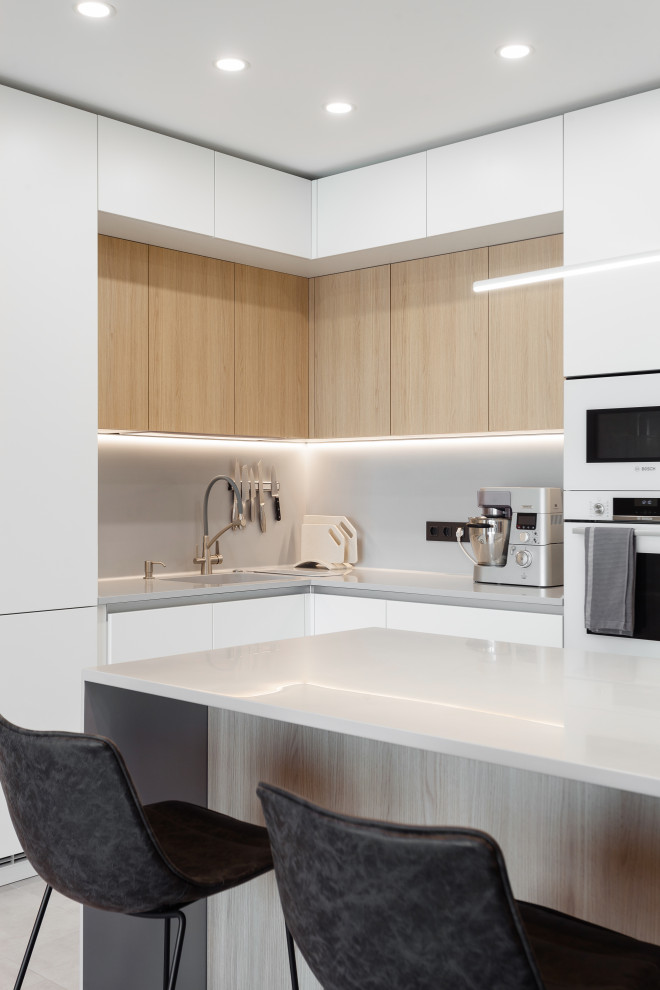 Mid-sized contemporary l-shaped open plan kitchen in Moscow with an undermount sink, flat-panel cabinets, white cabinets, solid surface benchtops, grey splashback, white appliances, porcelain floors, a peninsula, grey floor, grey benchtop and recessed.