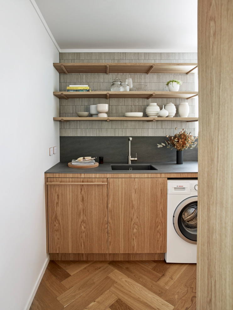 Inspiration for a small midcentury galley utility room in Auckland with an undermount sink, medium wood cabinets, tile benchtops, beige splashback, stone tile splashback, white walls, medium hardwood floors, a side-by-side washer and dryer, brown floor and grey benchtop.
