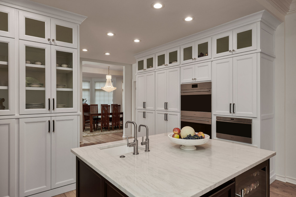 This is an example of an expansive transitional eat-in kitchen in Austin with a farmhouse sink, recessed-panel cabinets, white cabinets, quartz benchtops, grey splashback, marble splashback, black appliances, light hardwood floors, multiple islands, brown floor, white benchtop and coffered.