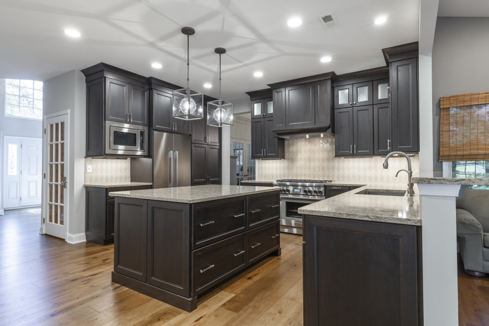 This is an example of a mid-sized transitional l-shaped eat-in kitchen in Philadelphia with an undermount sink, recessed-panel cabinets, black cabinets, quartz benchtops, beige splashback, porcelain splashback, stainless steel appliances, medium hardwood floors, with island, brown floor and beige benchtop.