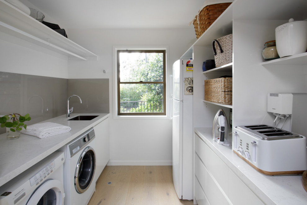 Design ideas for a small contemporary utility room in Brisbane with an undermount sink, white cabinets, quartz benchtops, metallic splashback, white walls, light hardwood floors, a side-by-side washer and dryer and white benchtop.