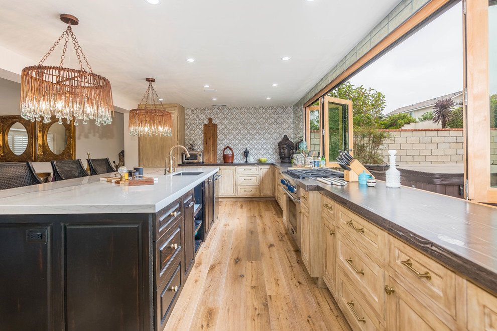 Photo of a large tropical l-shaped separate kitchen in Orange County with an undermount sink, recessed-panel cabinets, light wood cabinets, marble benchtops, metallic splashback, mirror splashback, panelled appliances, light hardwood floors, with island, brown floor and grey benchtop.
