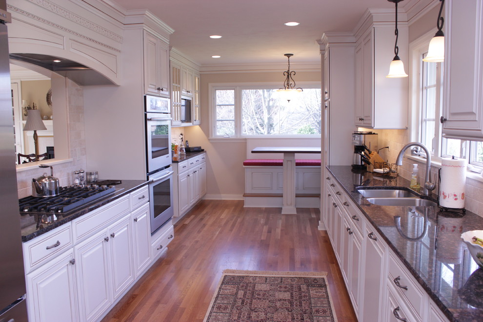 Large transitional galley medium tone wood floor and brown floor eat-in kitchen photo with a double-bowl sink, recessed-panel cabinets, white cabinets, granite countertops, beige backsplash, travertine backsplash, stainless steel appliances and no island