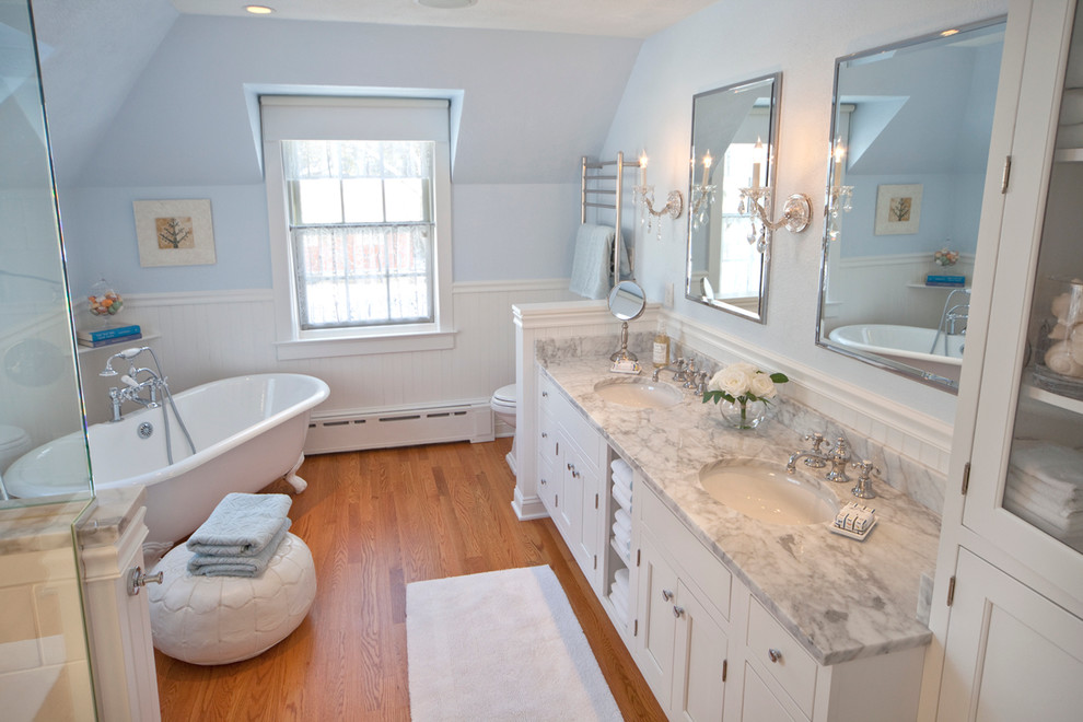 Example of a large classic master medium tone wood floor and brown floor claw-foot bathtub design with an undermount sink, a one-piece toilet, flat-panel cabinets, white cabinets, marble countertops, blue walls and a hinged shower door