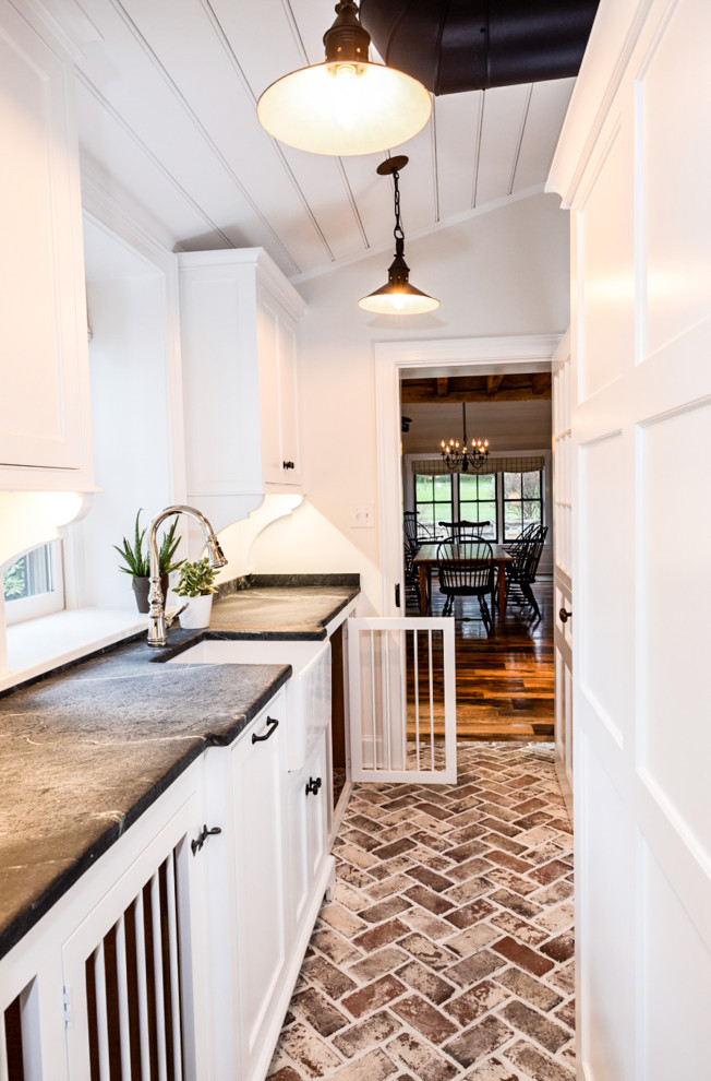 Inspiration for a small country galley utility room in Philadelphia with a farmhouse sink, beaded inset cabinets, white cabinets, soapstone benchtops, white walls, brick floors, a stacked washer and dryer, black benchtop and vaulted.