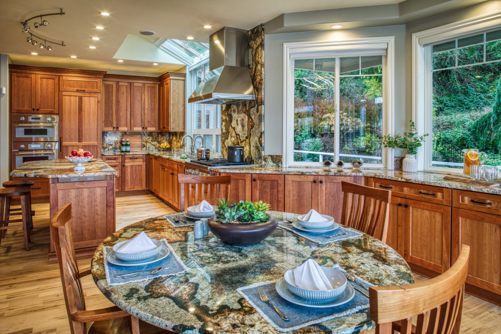 Photo of a large transitional l-shaped eat-in kitchen in Seattle with an undermount sink, recessed-panel cabinets, medium wood cabinets, granite benchtops, multi-coloured splashback, granite splashback, stainless steel appliances, light hardwood floors, with island and multi-coloured benchtop.
