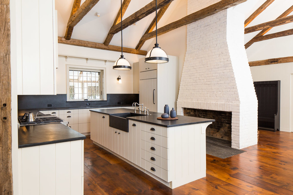 This is an example of a country l-shaped kitchen in Boston with a farmhouse sink, white cabinets, black splashback, medium hardwood floors, with island, brown floor and black benchtop.