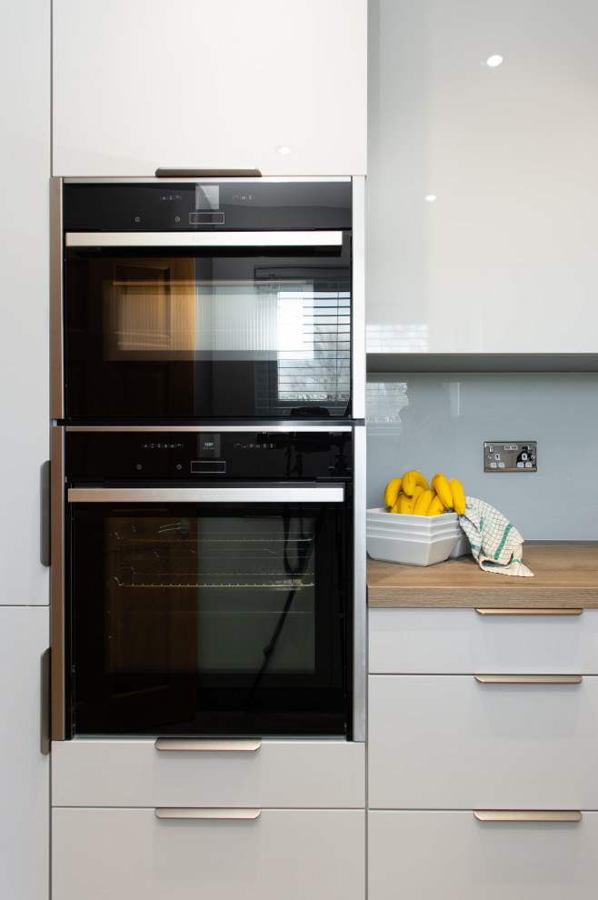 Inspiration for a small contemporary u-shaped enclosed kitchen in Cornwall with a built-in sink, flat-panel cabinets, grey cabinets, composite countertops, white splashback, glass sheet splashback, black appliances, light hardwood flooring, beige floors and beige worktops.