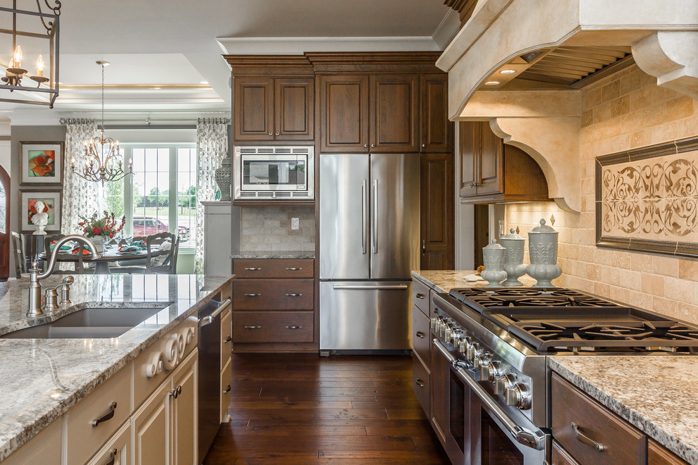This is an example of a mid-sized mediterranean l-shaped open plan kitchen in Louisville with an undermount sink, raised-panel cabinets, medium wood cabinets, granite benchtops, beige splashback, stone tile splashback, stainless steel appliances, medium hardwood floors and with island.