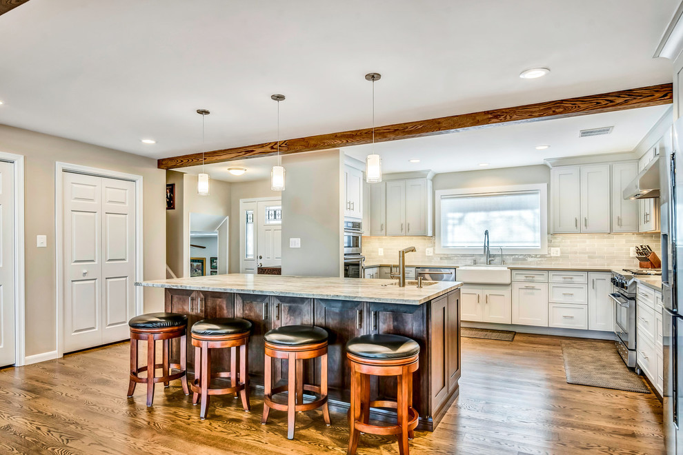 Example of a transitional medium tone wood floor and brown floor eat-in kitchen design in DC Metro with a farmhouse sink, shaker cabinets, white cabinets, granite countertops, white backsplash, subway tile backsplash, stainless steel appliances, an island and multicolored countertops