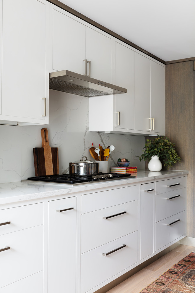 This is an example of a mid-sized contemporary single-wall open plan kitchen in New York with flat-panel cabinets, light wood cabinets, quartz benchtops, white splashback, marble splashback, stainless steel appliances, light hardwood floors, with island, beige floor and white benchtop.