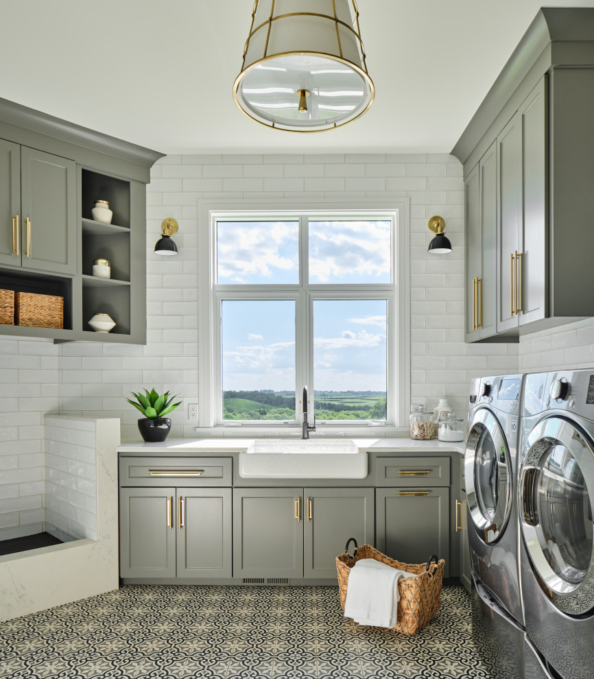Inspiration for a mid-sized transitional u-shaped dedicated laundry room in Omaha with a farmhouse sink, shaker cabinets, grey cabinets, quartz benchtops, white splashback, subway tile splashback, white walls, porcelain floors, a side-by-side washer and dryer, multi-coloured floor and grey benchtop.