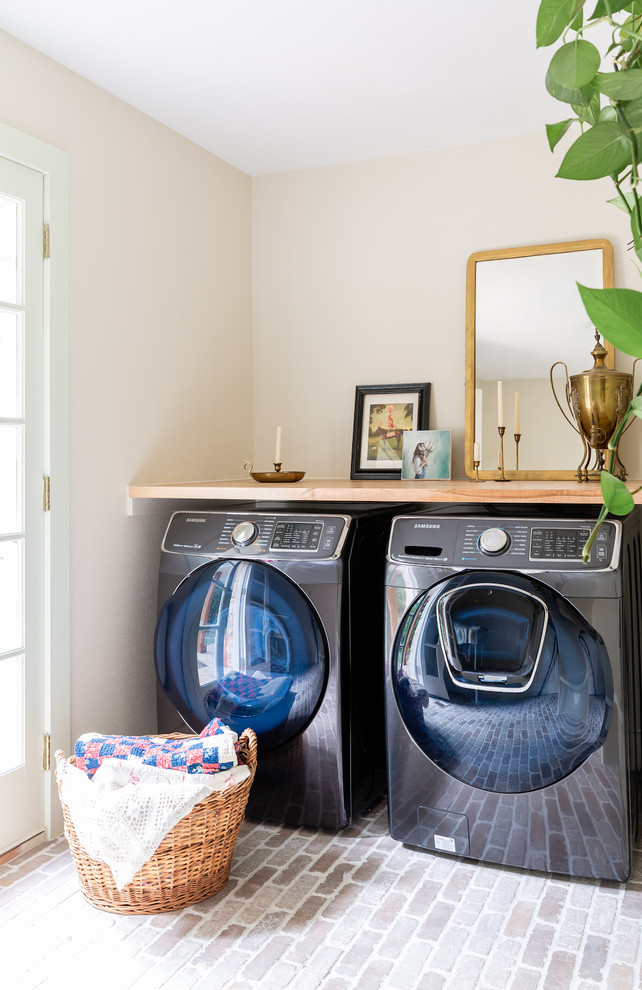 Photo of a mid-sized transitional dedicated laundry room in Dallas with wood benchtops, brick floors, a side-by-side washer and dryer, brown benchtop and beige walls.