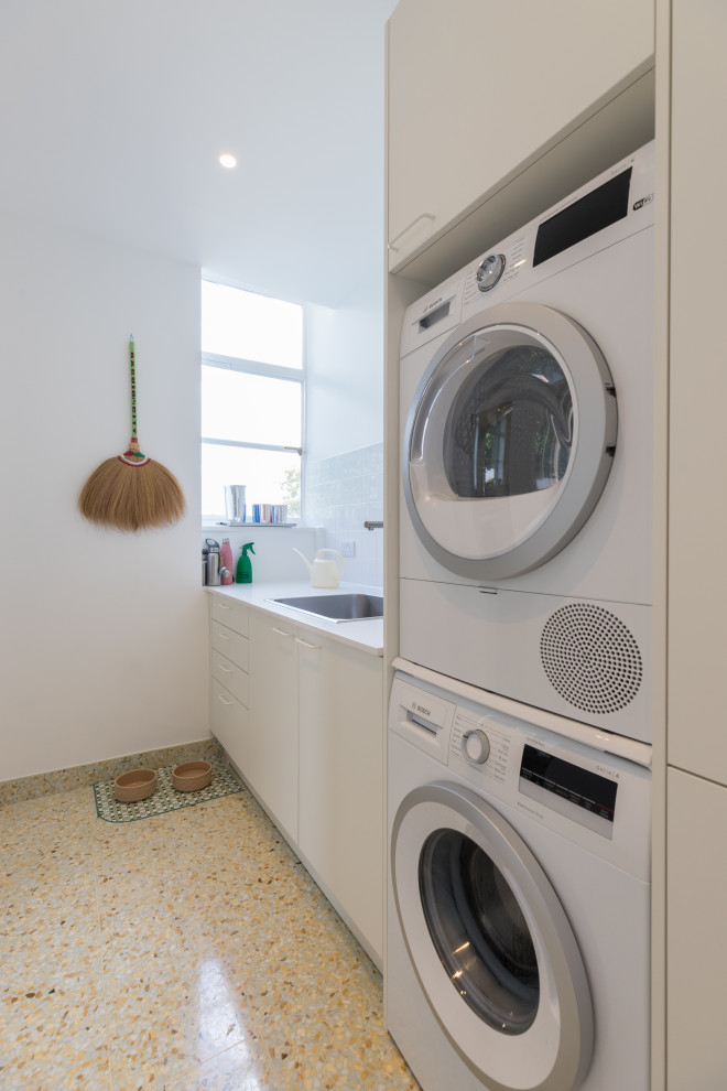 Design ideas for a large contemporary l-shaped dedicated laundry room in London with a drop-in sink, flat-panel cabinets, white cabinets, solid surface benchtops, white splashback, ceramic splashback, white walls, porcelain floors, a stacked washer and dryer, green floor and white benchtop.