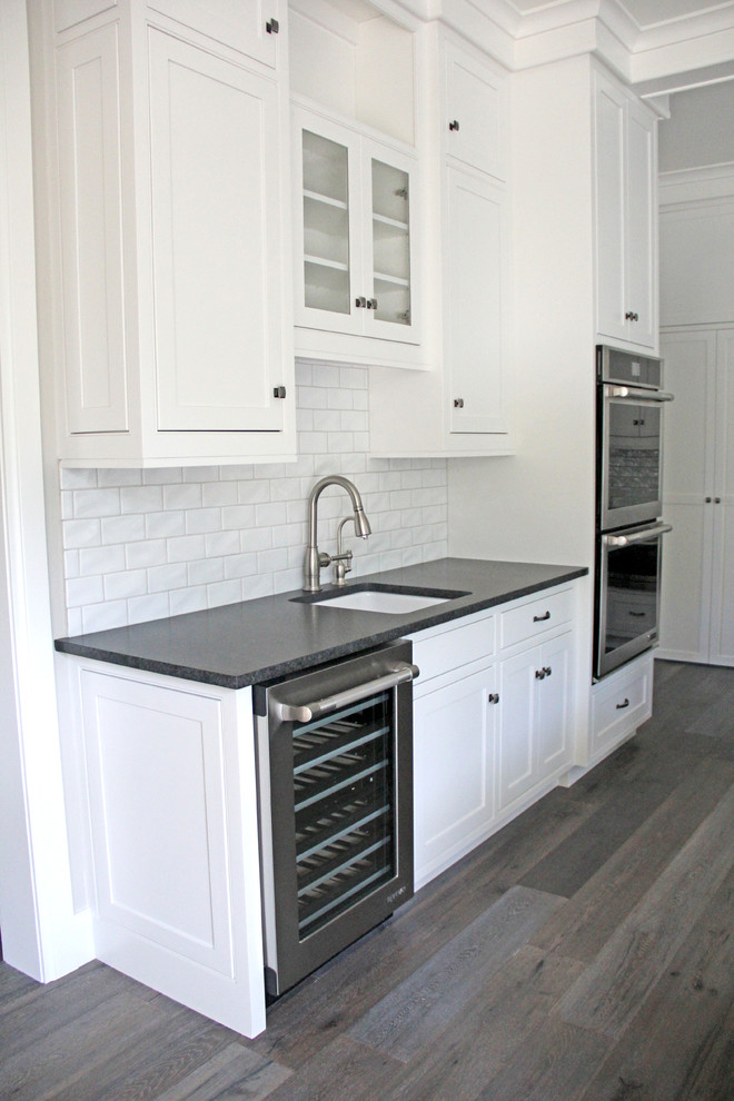 Large single-wall light wood floor and brown floor eat-in kitchen photo with an undermount sink, recessed-panel cabinets, white cabinets, granite countertops, white backsplash, subway tile backsplash, stainless steel appliances and an island