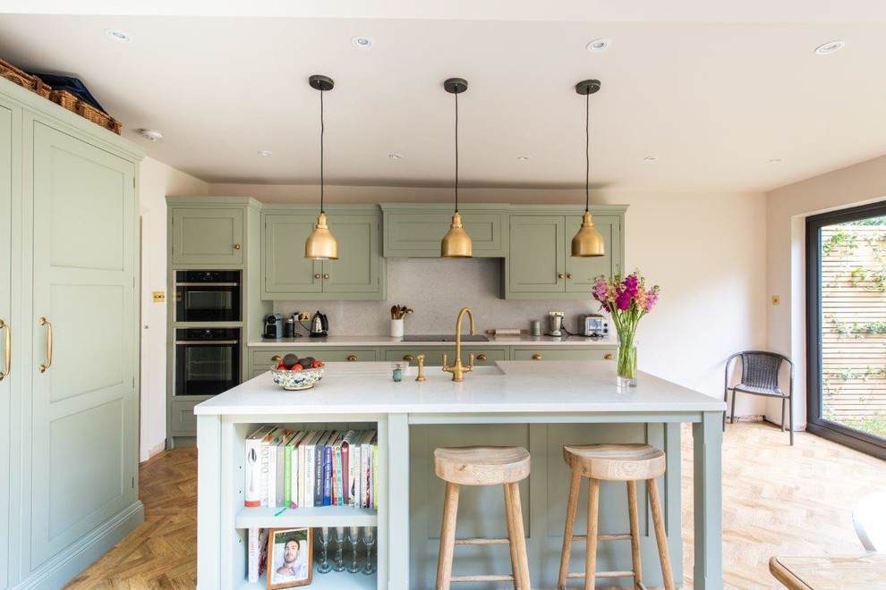 This is an example of a mid-sized transitional kitchen in Kent with a farmhouse sink, green cabinets, white splashback, marble splashback, light hardwood floors, with island, beige floor, white benchtop, shaker cabinets and black appliances.