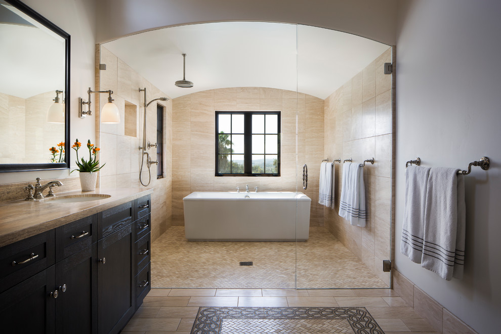 Photo of a large mediterranean master wet room bathroom in Santa Barbara with dark wood cabinets, a freestanding tub, beige tile, travertine, ceramic floors, an undermount sink, beige floor, a hinged shower door, recessed-panel cabinets, white walls and brown benchtops.