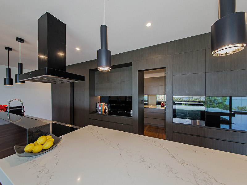 Photo of a large modern galley eat-in kitchen in Brisbane with with island, a drop-in sink, dark wood cabinets, stainless steel benchtops, black splashback, glass sheet splashback, black appliances and bamboo floors.
