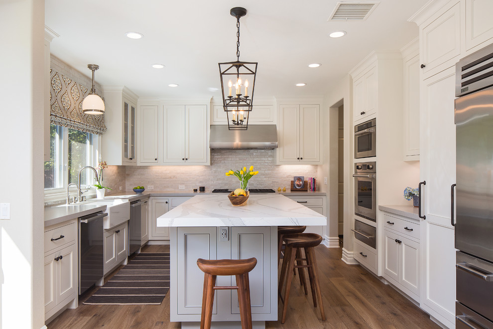 This is an example of a mid-sized transitional u-shaped kitchen in Los Angeles with with island, a farmhouse sink, recessed-panel cabinets, white cabinets, marble benchtops, stainless steel appliances, dark hardwood floors, brown floor, white benchtop, grey splashback and subway tile splashback.