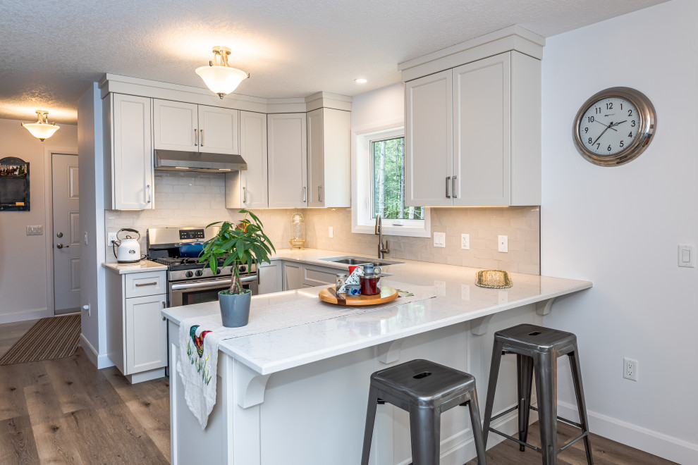This is an example of a transitional u-shaped kitchen in Toronto with an undermount sink, white cabinets, quartz benchtops, white splashback, ceramic splashback, stainless steel appliances, vinyl floors, with island, brown floor and white benchtop.