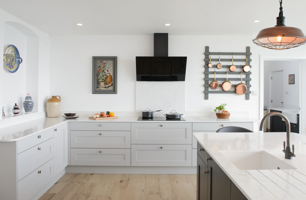This is an example of a large classic u-shaped kitchen/diner in Cornwall with a belfast sink, shaker cabinets, grey cabinets, black appliances, light hardwood flooring, an island, beige floors and white worktops.