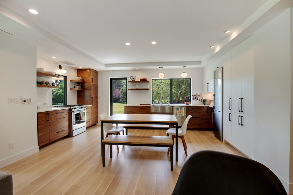 This is an example of a mid-sized modern l-shaped eat-in kitchen in Denver with a farmhouse sink, flat-panel cabinets, dark wood cabinets, quartz benchtops, white splashback, ceramic splashback, stainless steel appliances, light hardwood floors, no island, brown floor and white benchtop.